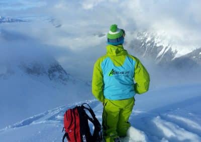 A hiker in bright green winter gear stands on a snowy mountain, overlooking a cloud-covered valley with a red backpack beside them.
