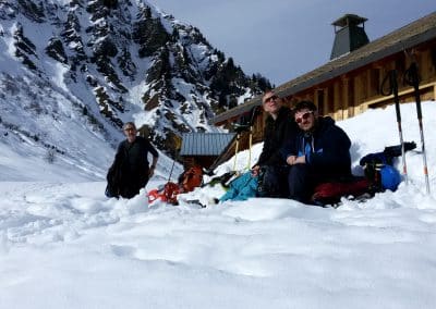 Three individuals resting in deep snow near a mountain cabin, surrounded by snow-covered terrain and winter gear.